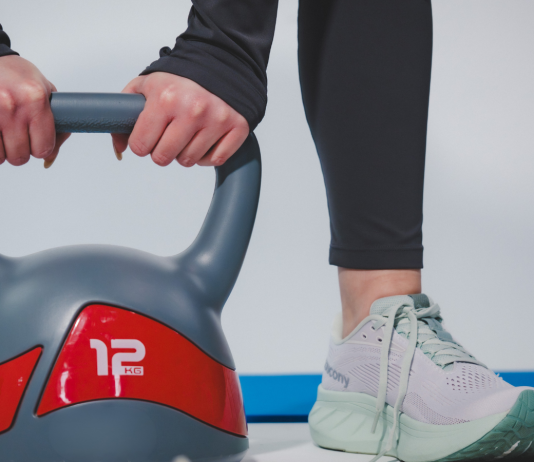 Donna che solleva un kettlebell da 12 kg durante un allenamento fitness indoor con abbigliamento sportivo e scarpe da training