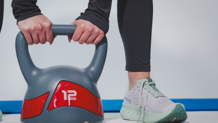 Donna che solleva un kettlebell da 12 kg durante un allenamento fitness indoor con abbigliamento sportivo e scarpe da training